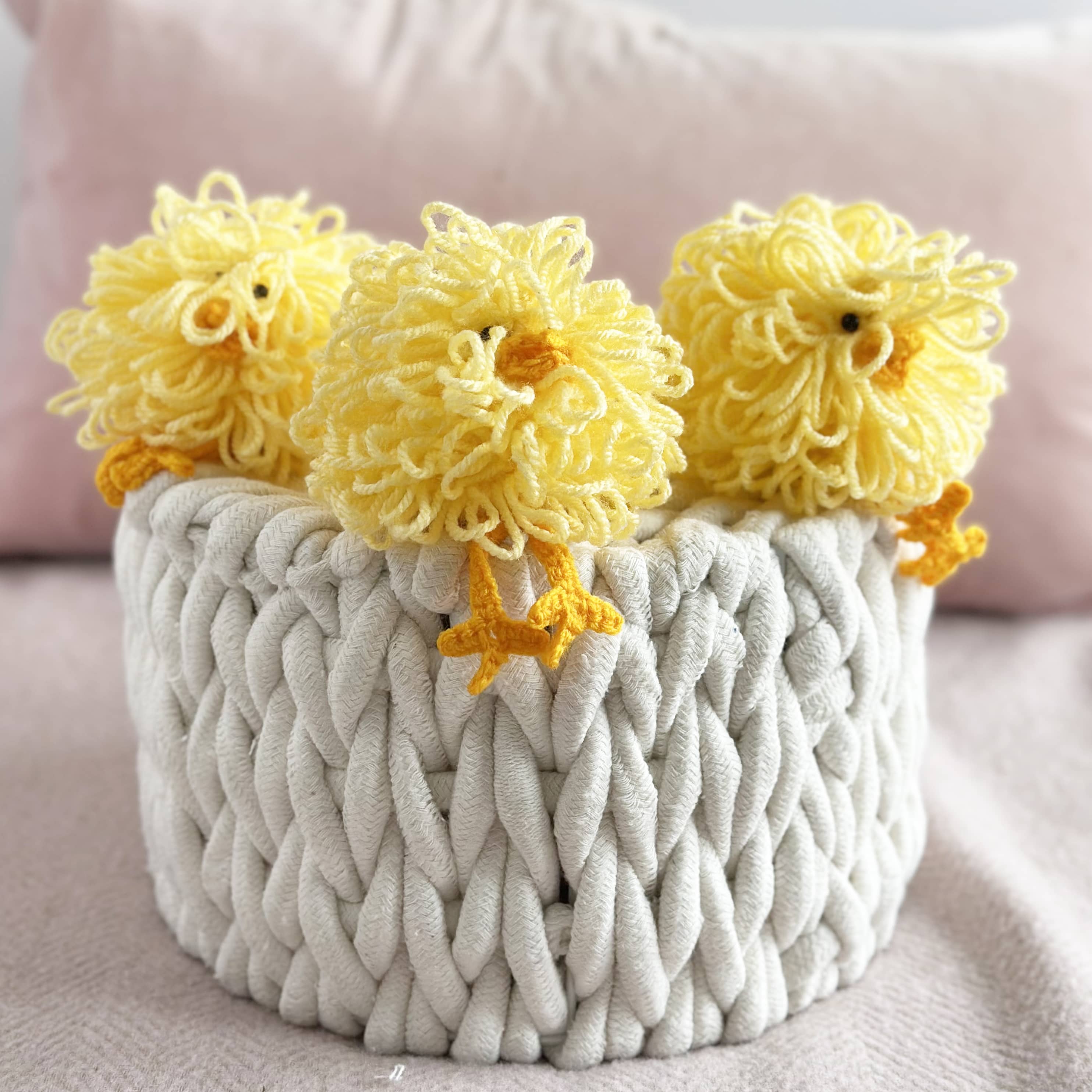 Three yellow knitted chicks in a textured white basket on a soft pink background