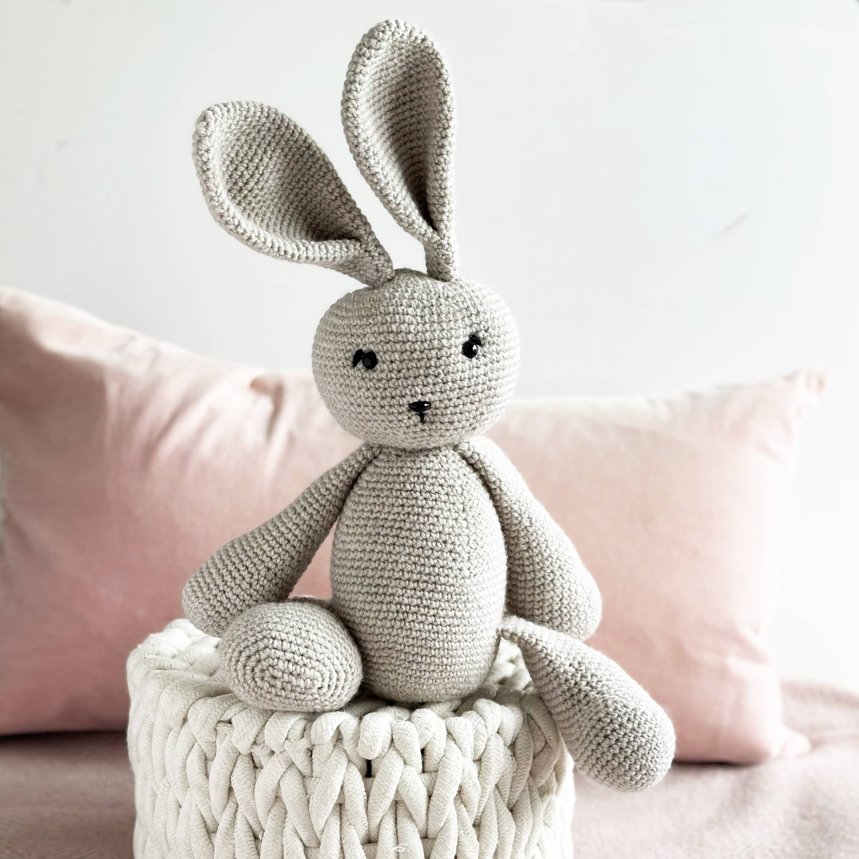 Crocheted gray bunny toy on a white woven basket with pink cushions in the background