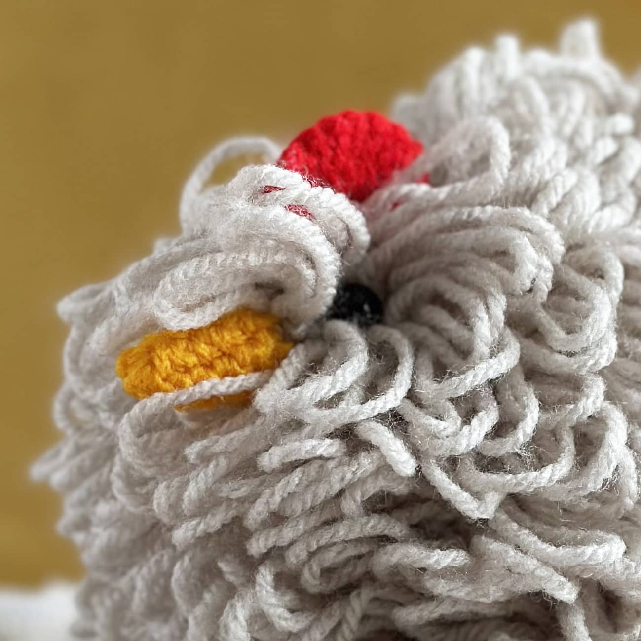 Close-up of a knitted chicken with red head and yellow beak on a beige background
