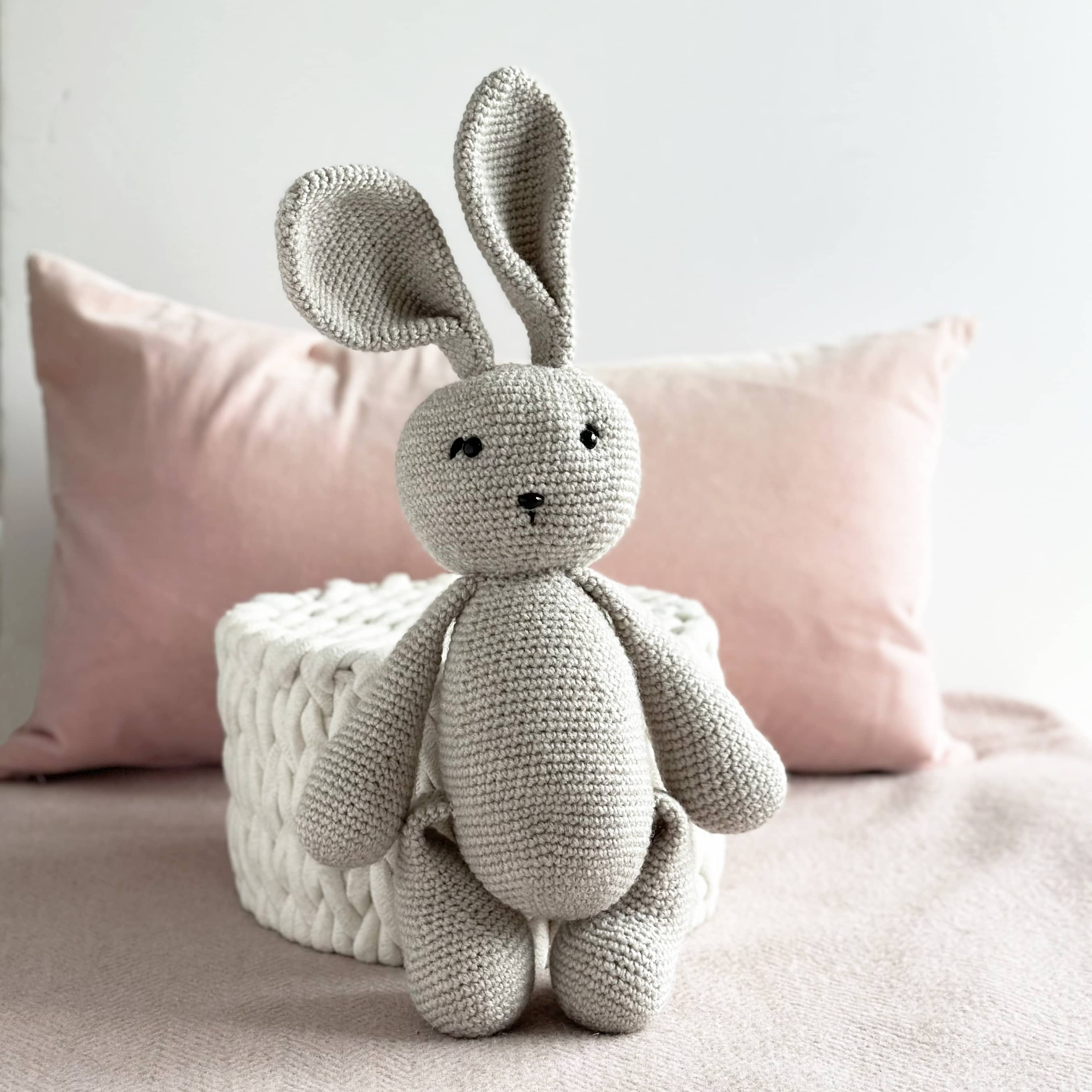 Crocheted bunny toy on a bed with pink pillows and a textured white box.