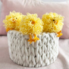 Three yellow knitted chicks in a textured white basket on a soft pink background